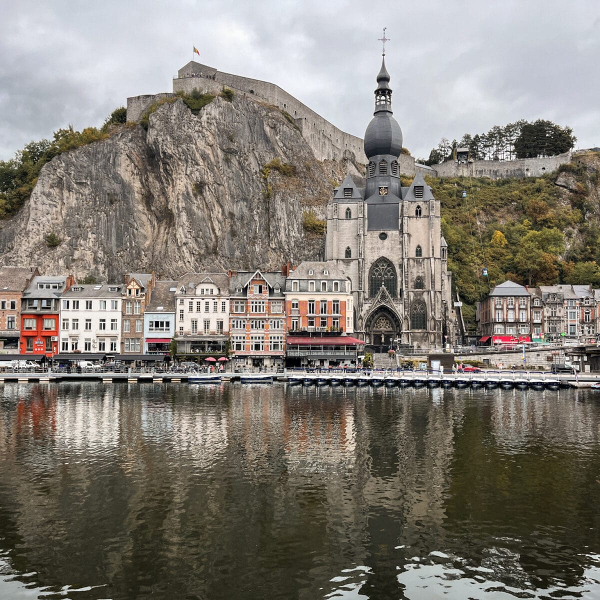 Colorful buildings on a riverside next to a gothic style church with a stone mountain in the background.